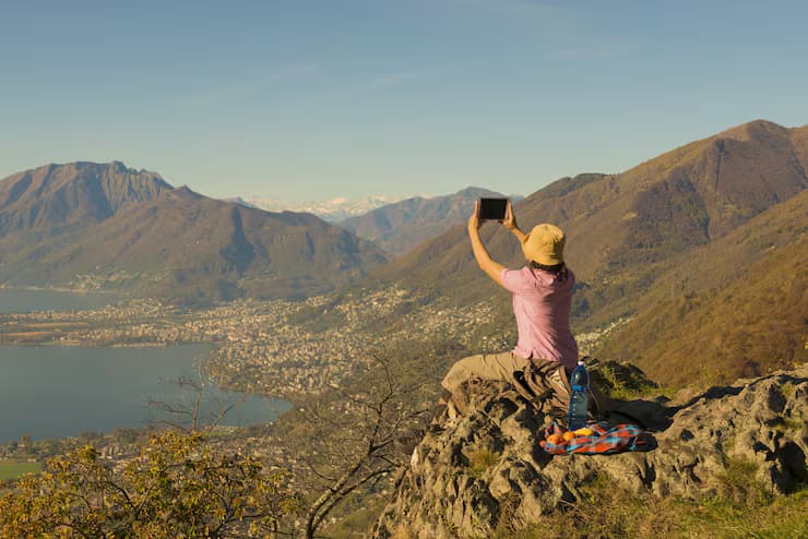 Traveler sitting on a cliff edge taking a photo of a large lake and mountains with a tablet