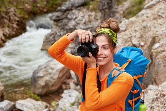 Female hiker in an orange sweater photographing a mountain stream with a DSLR camera