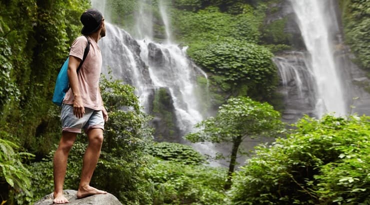 Traveler standing on a rock gazing up at a majestic waterfall in a lush forest
